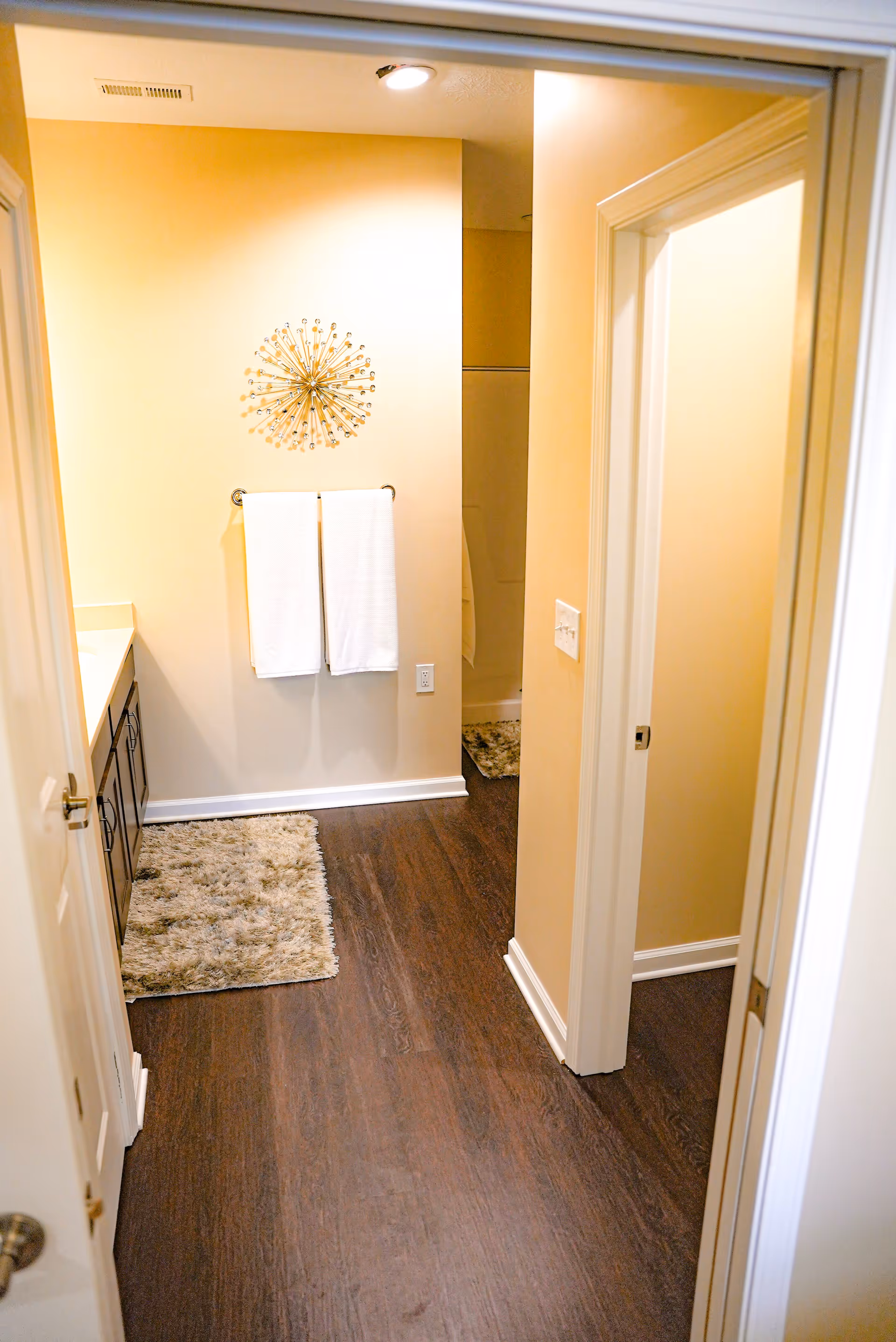 View into a bathroom with dark wood flooring, a beige wall with a decorative metal wall art above two white towels hanging on a towel rack, a countertop with cabinets on the left, and a glimpse of a shower area in the background.