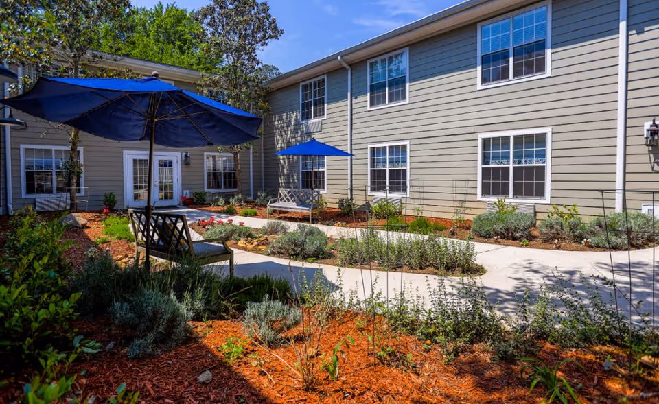 Outdoor courtyard area at Addington Place of Roswell featuring a paved walkway, garden beds with various plants, benches, and blue umbrellas providing shade. The building exterior is visible with multiple windows and light gray siding under a clear blue sky.
