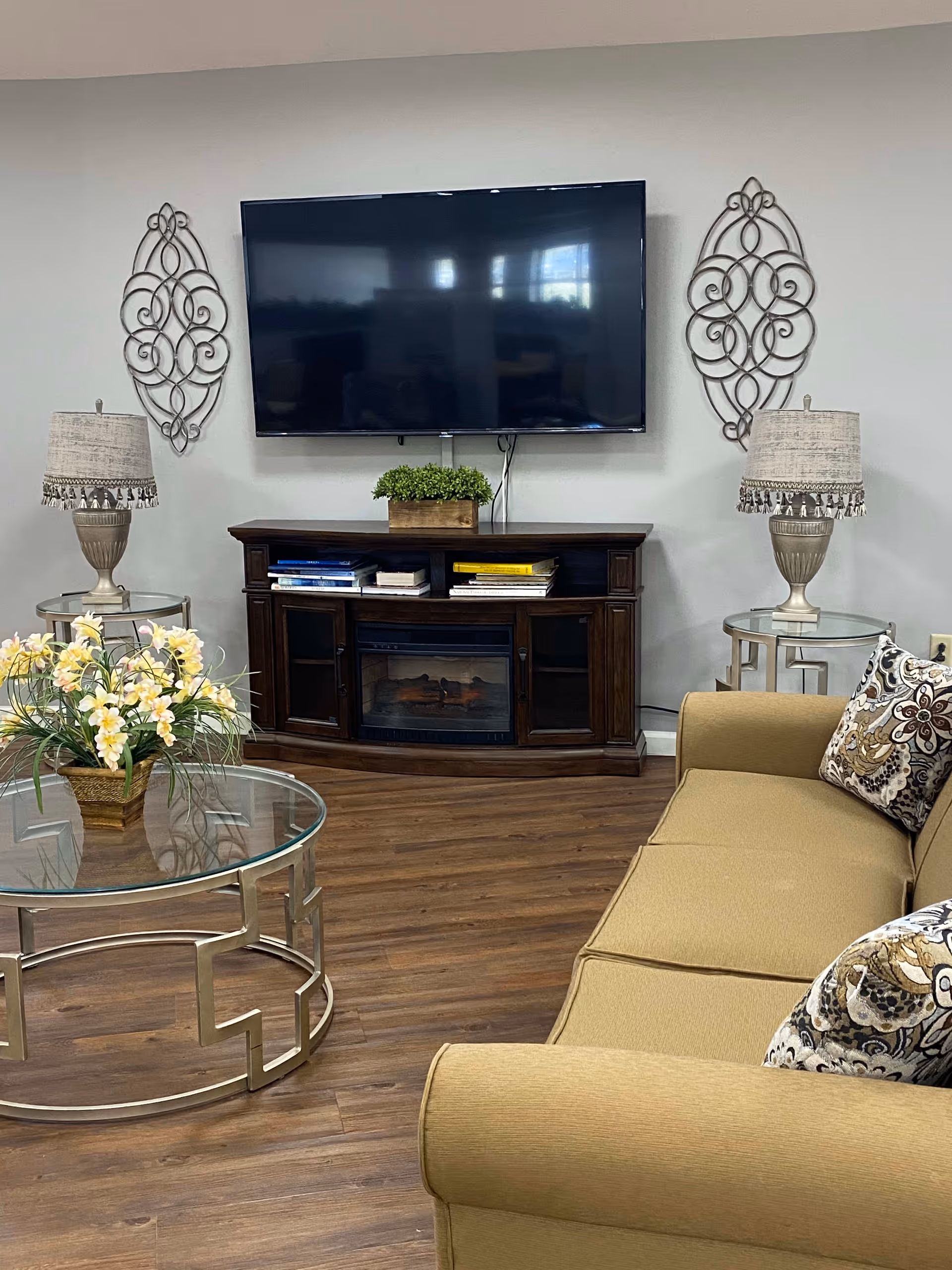 A cozy living room with a beige sofa adorned with patterned cushions, a glass coffee table with a floral arrangement, two side tables each with a lamp, a dark wooden TV stand with books and a small plant on top, and a large flat-screen TV mounted on the wall. Decorative metal wall art pieces are hung on either side of the TV.