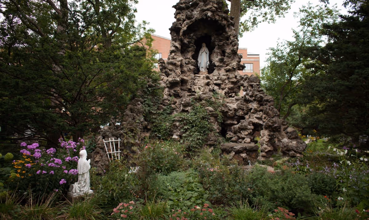 Outdoor garden area with lush greenery and flowering plants surrounding a large rock grotto. The grotto features a statue of the Virgin Mary in a niche near the top, and another statue of a kneeling figure in prayer is positioned among the flowers. Trees and a brick building are visible in the background.