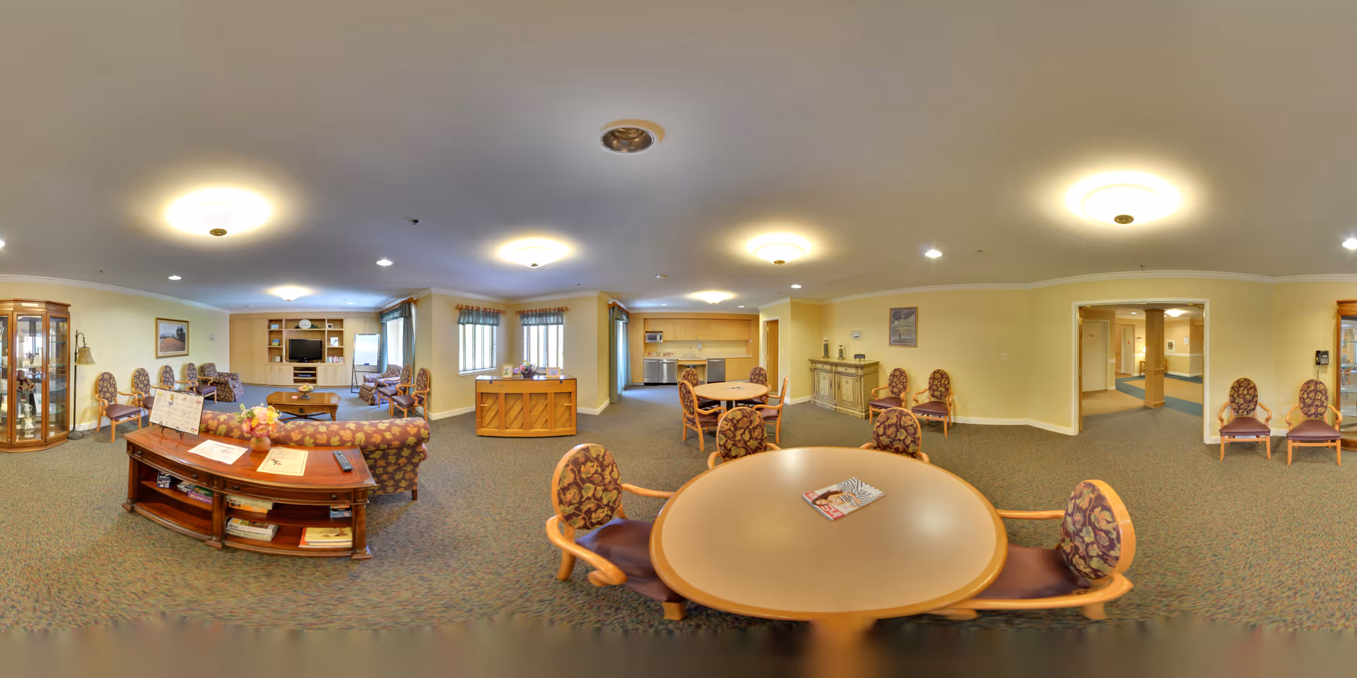 A spacious, well-lit common area in a senior living community featuring multiple round tables with patterned chairs, a piano, a wooden cabinet with books and decorations, and a seating area with sofas and armchairs. The room has beige walls, carpeted floors, and several ceiling lights. There are windows with curtains and a hallway leading to other rooms.