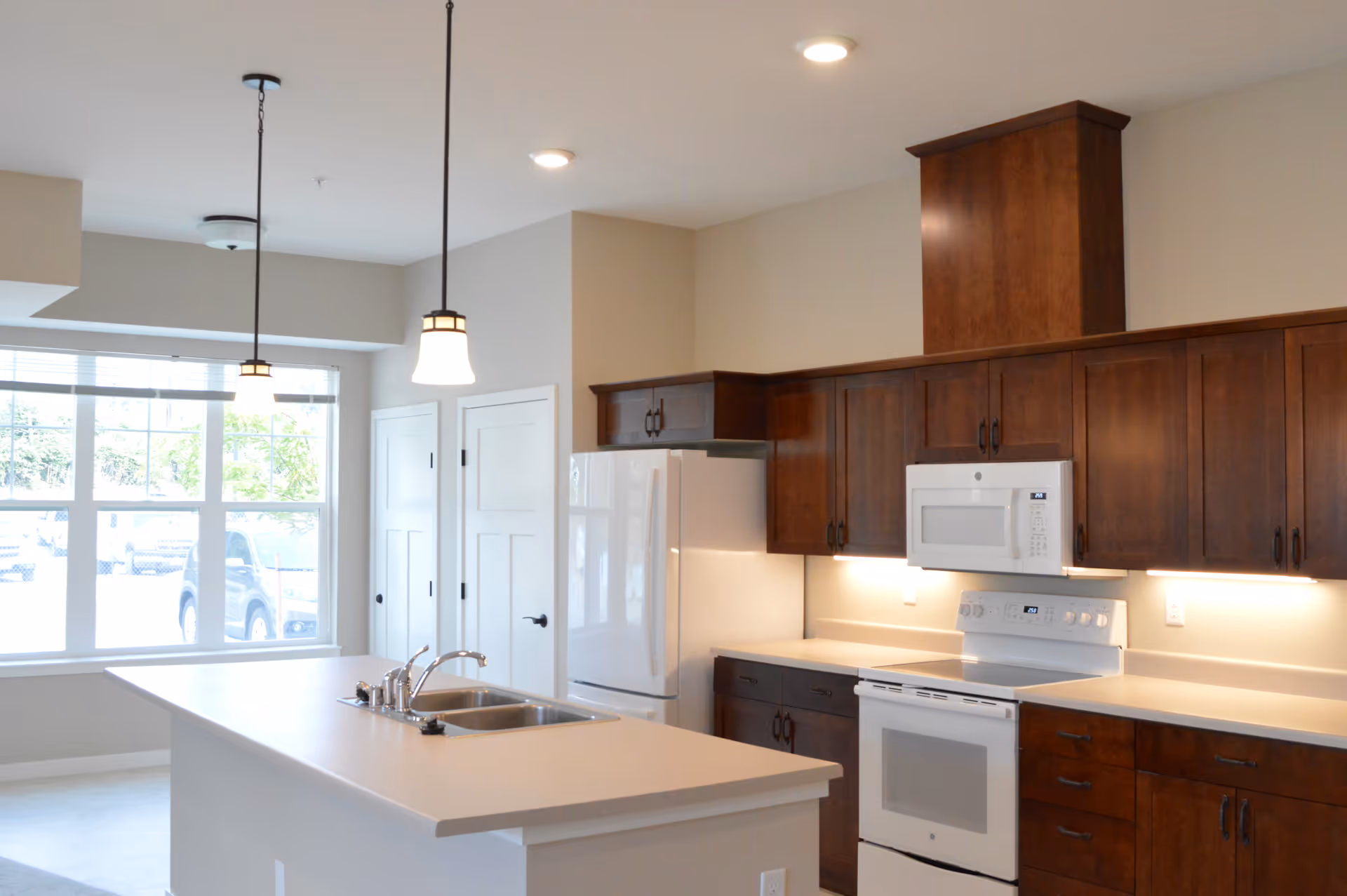 Bright kitchen interior with dark wooden cabinets, white countertops, a white refrigerator, stove, and microwave. There is a kitchen island with a double sink and two pendant lights hanging above it. Large windows allow natural light to fill the space.