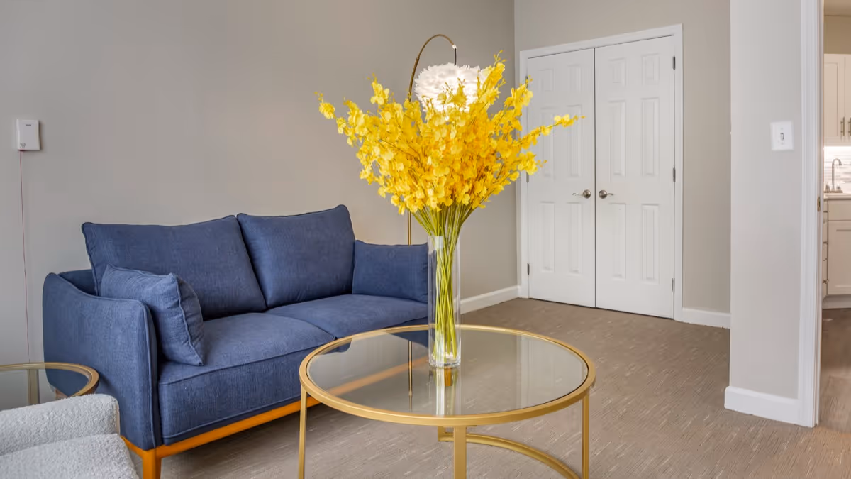 A modern living room with a blue sofa, a glass coffee table with a gold frame, and a tall vase filled with bright yellow flowers. The room has beige walls, a beige carpet, a white double-door closet, and a glimpse of a kitchen area to the right.
