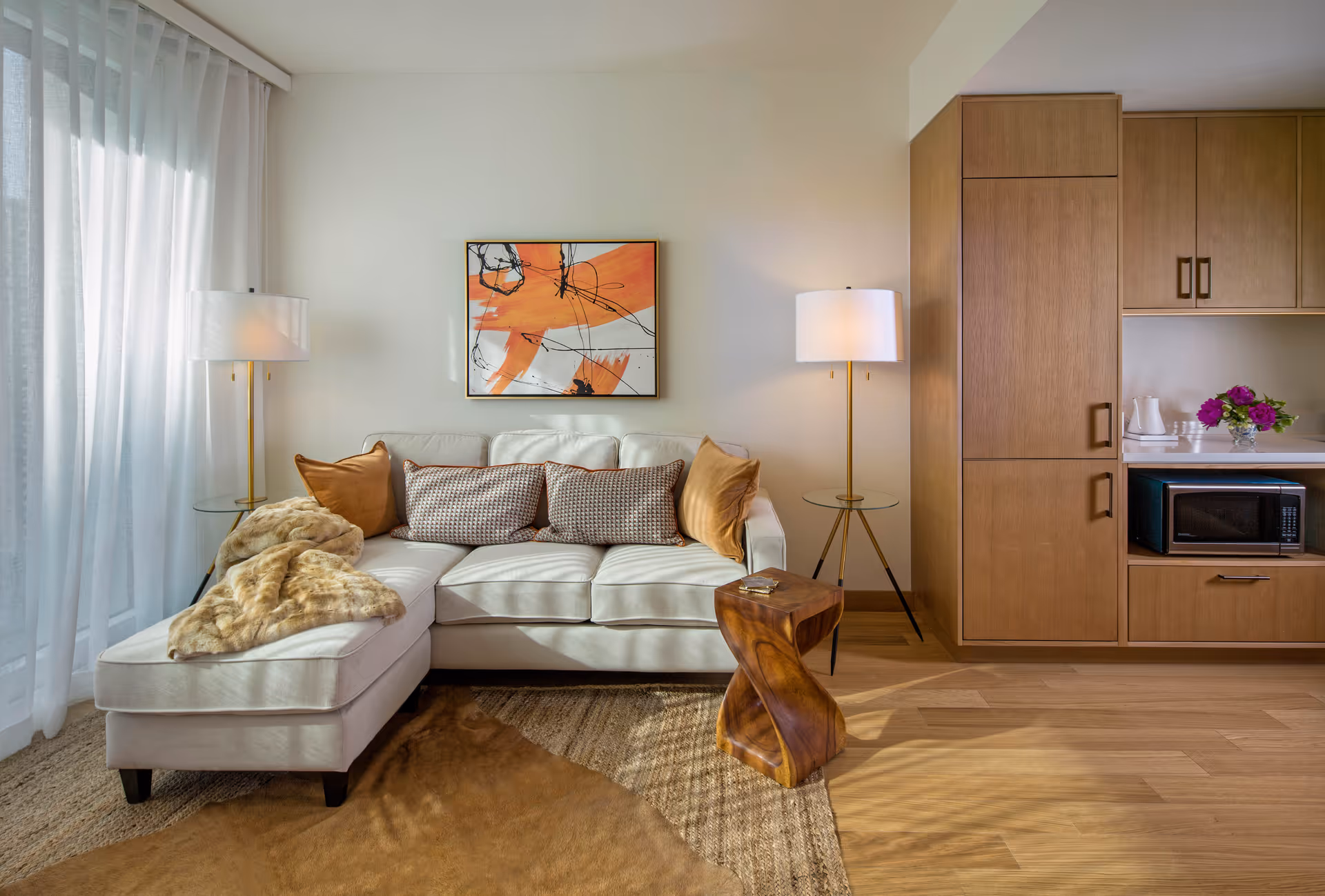 A cozy living room area with a white sectional sofa adorned with brown and patterned pillows, a soft fur throw, two modern floor lamps with white shades on either side, a small wooden side table, and a wooden cabinet unit with a microwave and a vase of flowers. A large abstract painting with orange and black strokes hangs on the wall above the sofa. Light filters through sheer white curtains on the left side.