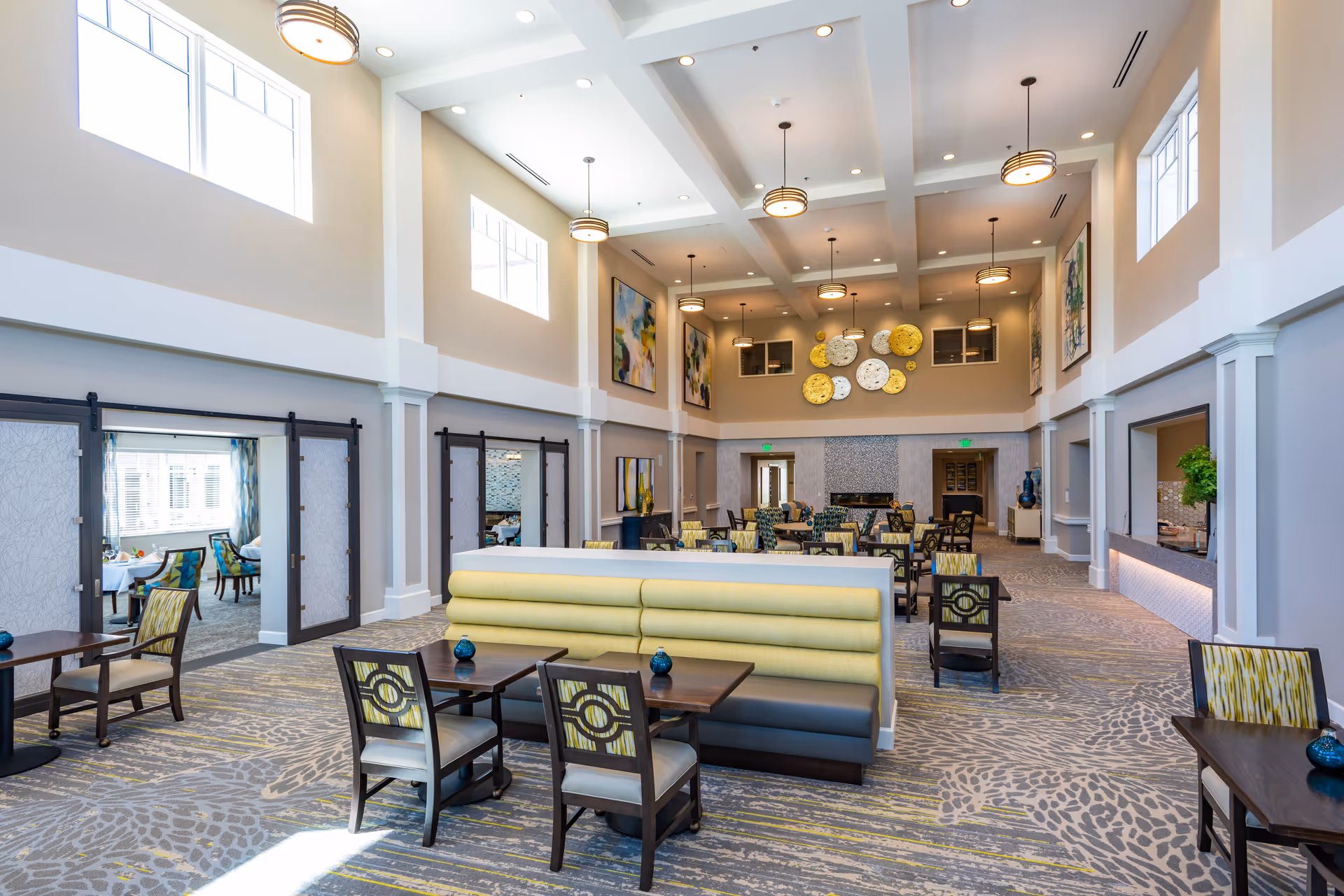 Spacious and well-lit dining area in a senior living facility with high ceilings, modern pendant lights, patterned carpet, and a mix of booth and chair seating. The room features large windows, colorful abstract wall art, and a serving counter on the right side.