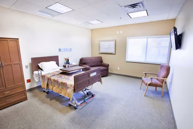 A senior care facility bedroom with a hospital-style bed covered with a floral blanket, a wooden wardrobe, a small couch, a single armchair, a wall-mounted TV, and a window with blinds letting in natural light.