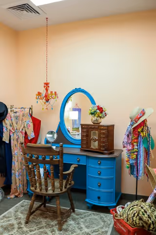 A cozy dressing area featuring a vintage wooden chair in front of a bright blue vanity with an oval mirror. The vanity holds a small wooden jewelry box and a vase with colorful flowers. To the left, a clothing rack displays various dresses, and to the right, a mannequin is adorned with hats, scarves, and leis. A decorative chandelier hangs from the ceiling, and a patterned rug covers the floor.