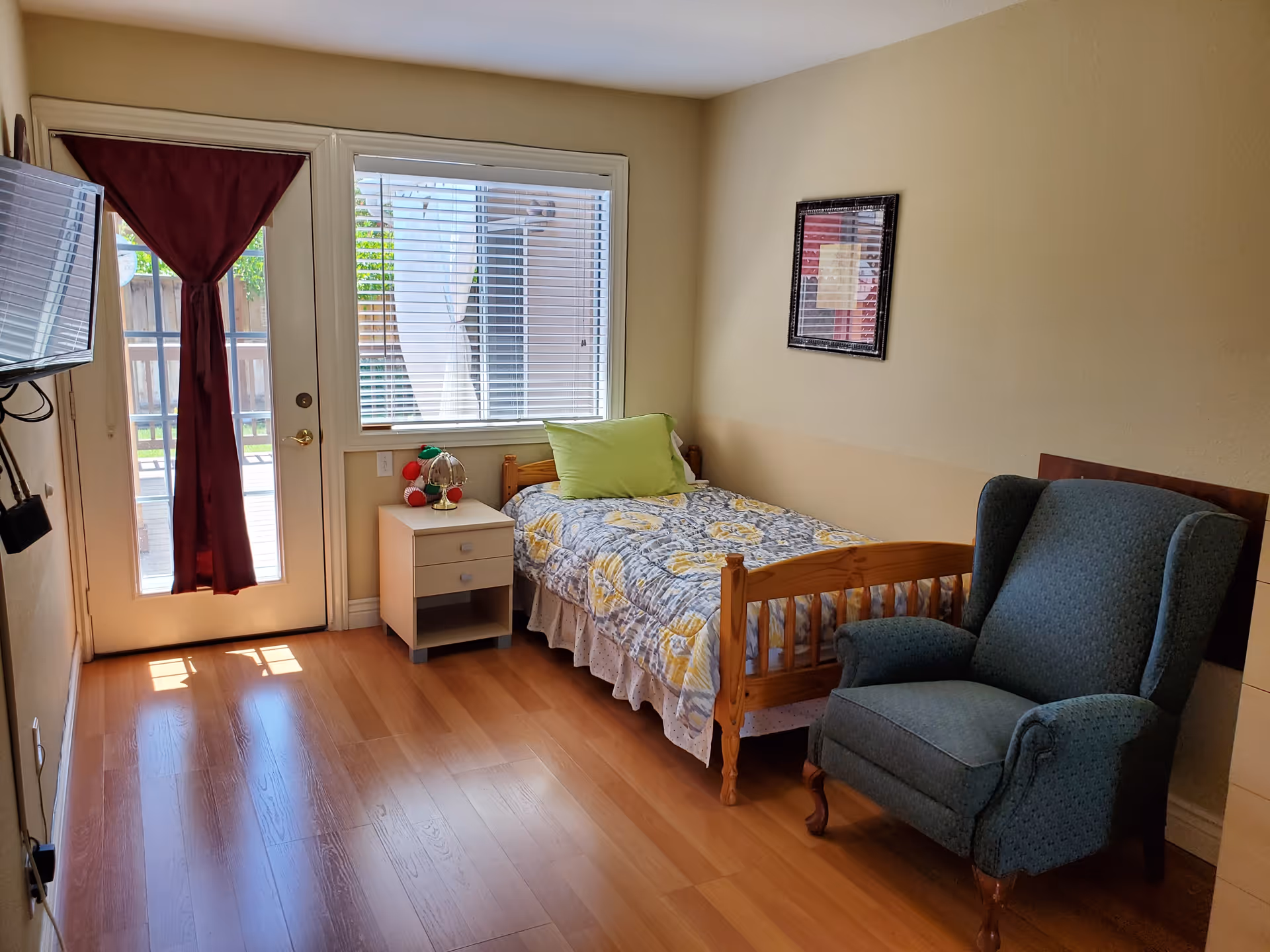 A small, cozy bedroom with a single wooden bed covered with a floral quilt and a green pillow. Next to the bed is a white nightstand with a decorative lamp and a small plush toy. A large window with white blinds and a door with a maroon curtain allow natural light into the room. There is a comfortable blue armchair near the bed, and a flat-screen TV mounted on the wall opposite the bed. The floor is wooden, and a framed picture hangs on the beige wall.