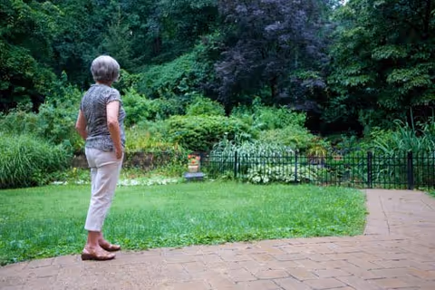 An elderly woman standing on a paved patio looking out towards a lush green garden with dense trees and shrubs, surrounded by a black metal fence.