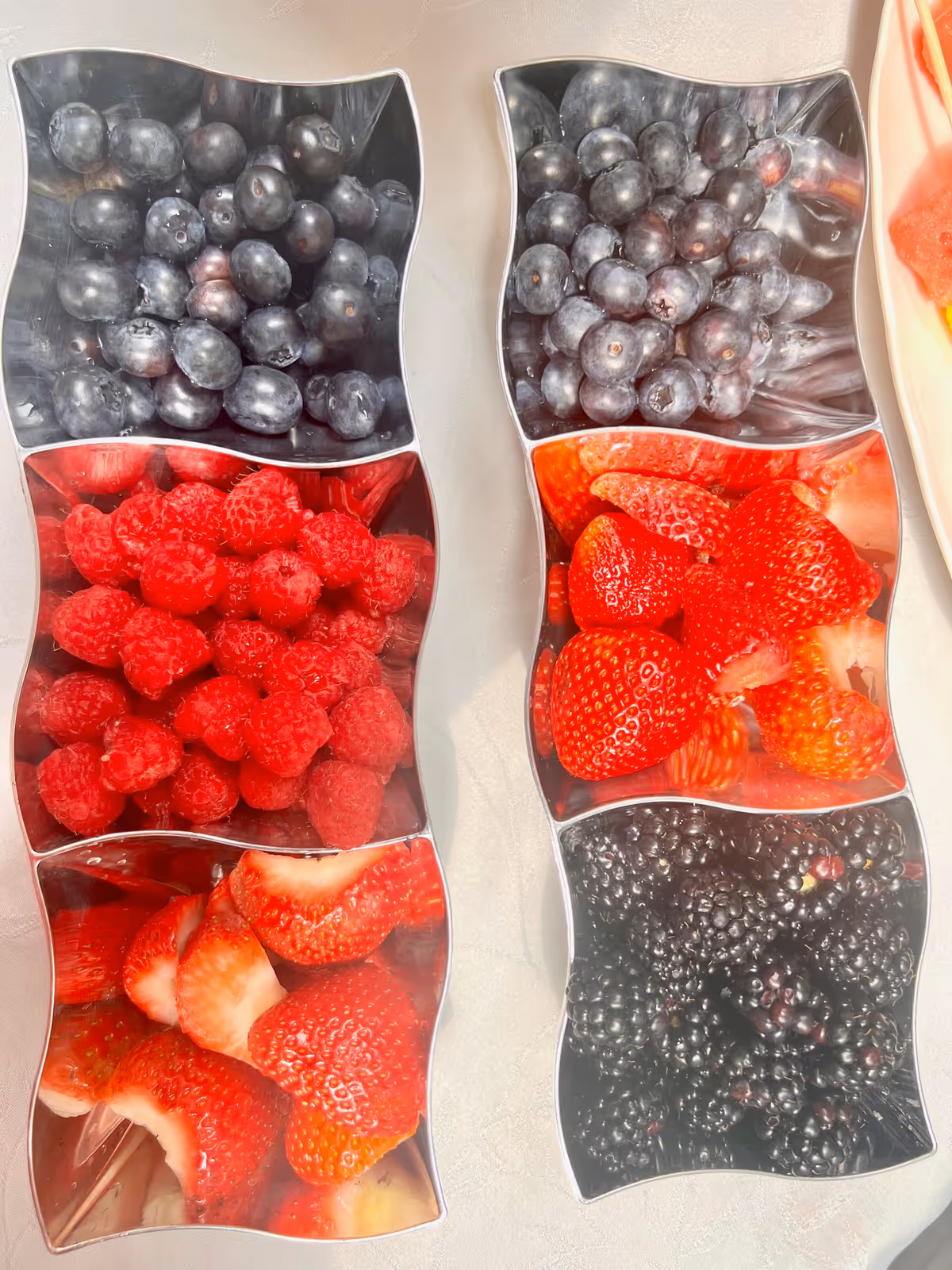 Two silver divided trays each containing three sections filled with fresh berries. The left tray has blueberries, raspberries, and sliced strawberries. The right tray has blueberries, whole strawberries, and blackberries.
