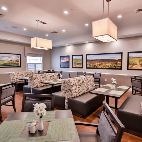 A dining room with several tables and cushioned booth seating. The tables have green placemats and small white flower vases. The walls are decorated with framed landscape paintings, and two large rectangular pendant lights hang from the ceiling.