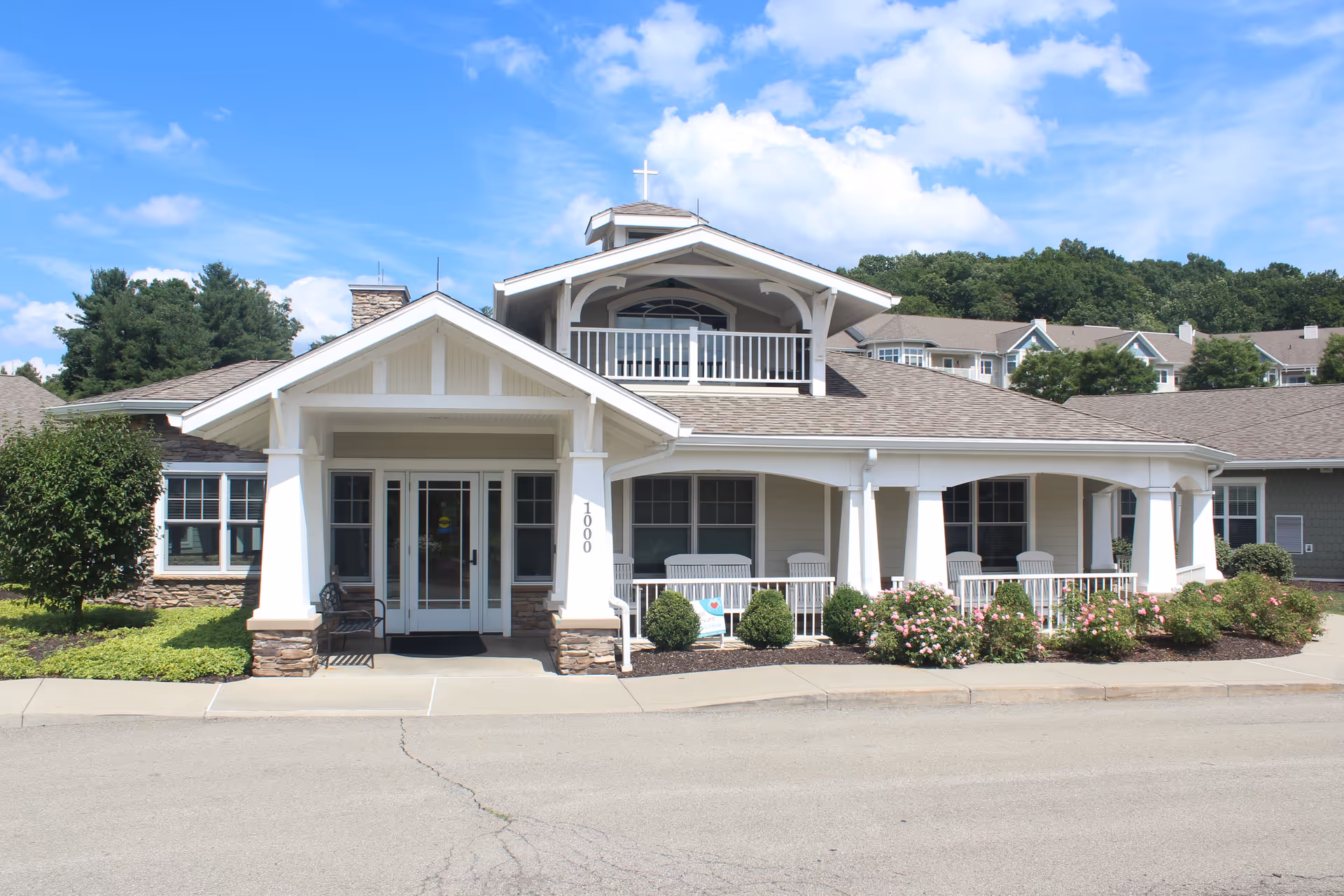 Front exterior view of St. John Community building with a covered entrance, white pillars, stone accents, a balcony with a cross on top, and landscaped bushes and flowers under a blue sky with some clouds.