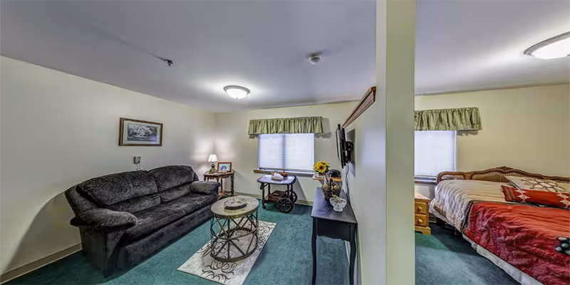 Interior view of a senior living facility room divided by a partial wall. On the left side, there is a living area with a dark sofa, a round coffee table on a small rug, a side table with a lamp, and a window with a green valance. On the right side, there is a bedroom area with a bed covered in a red and beige blanket, a wooden nightstand, and a window with a matching green valance. A TV is mounted on the wall facing the living area.