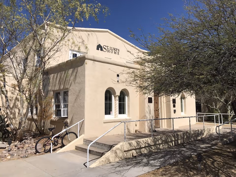 Front facade of a beige stucco building labeled "St. Luke's Home" with a wheelchair ramp, steps, a parked bicycle, and surrounding trees.