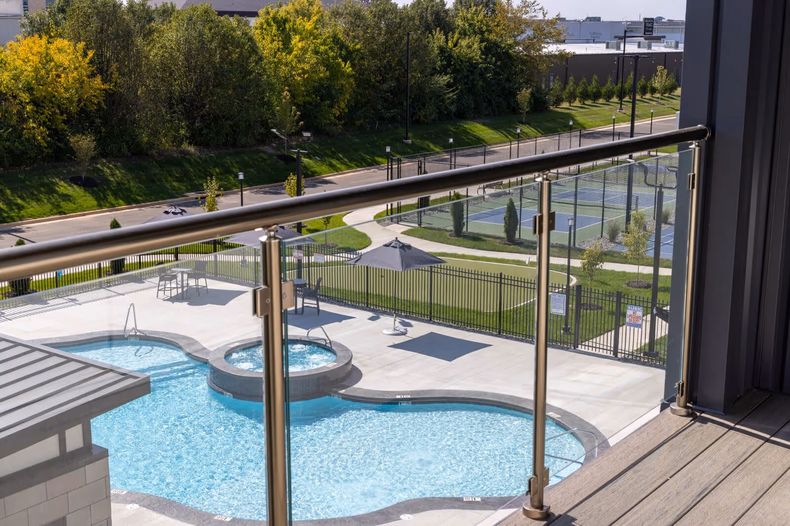 View from a balcony over a swimming pool and hot tub with adjacent tennis courts and landscaped greenery.