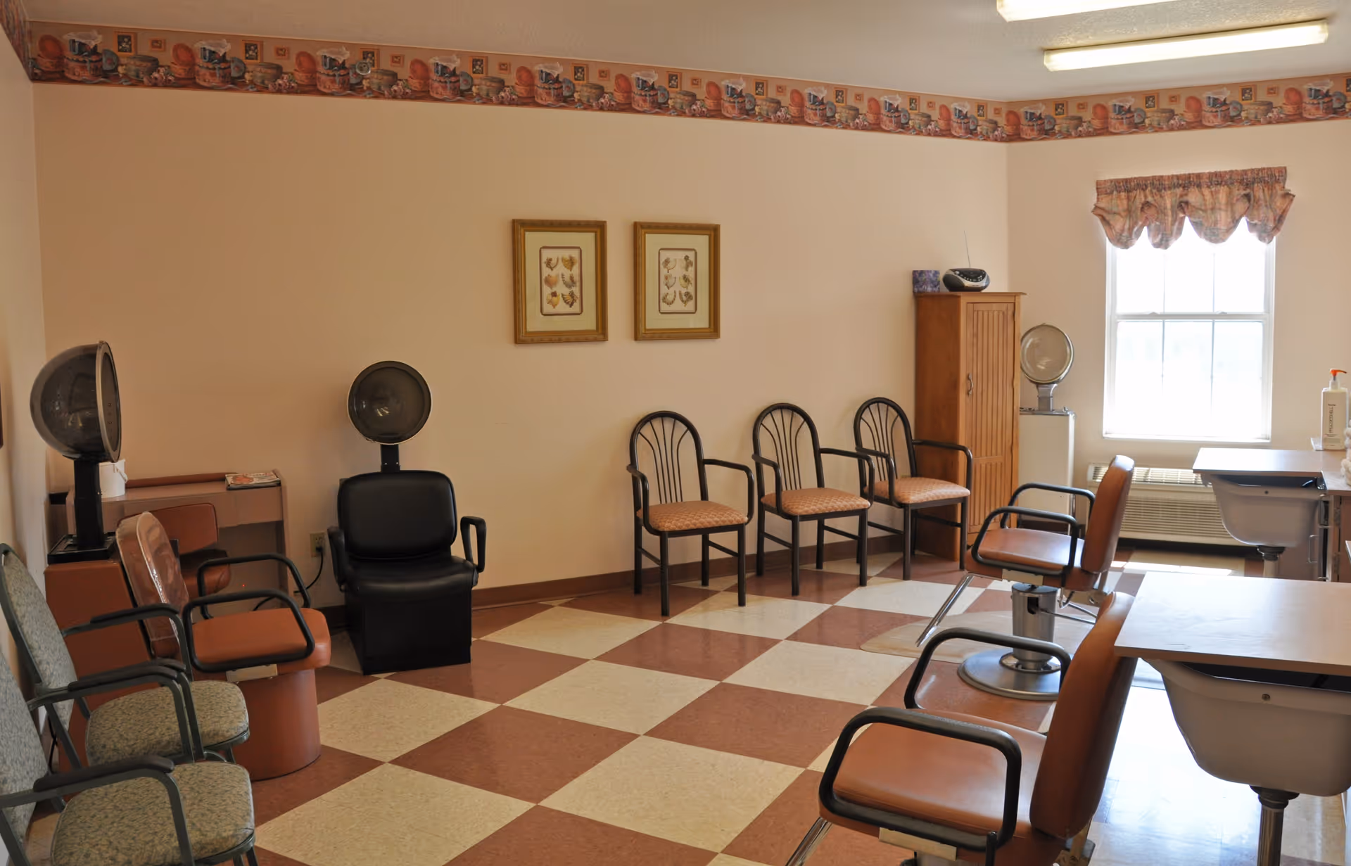 Interior of a hair salon area in a senior living facility with several salon chairs, hair dryers, and waiting chairs arranged along the walls. The room has a checkered floor, light-colored walls with a decorative border near the ceiling, two framed pictures, a window with a valance, and a wooden cabinet.