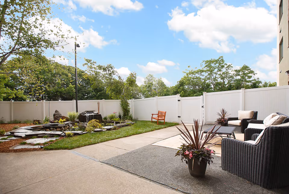 Sunny fenced courtyard with patio furniture, potted plants, a small pond feature, and a wooden bench.