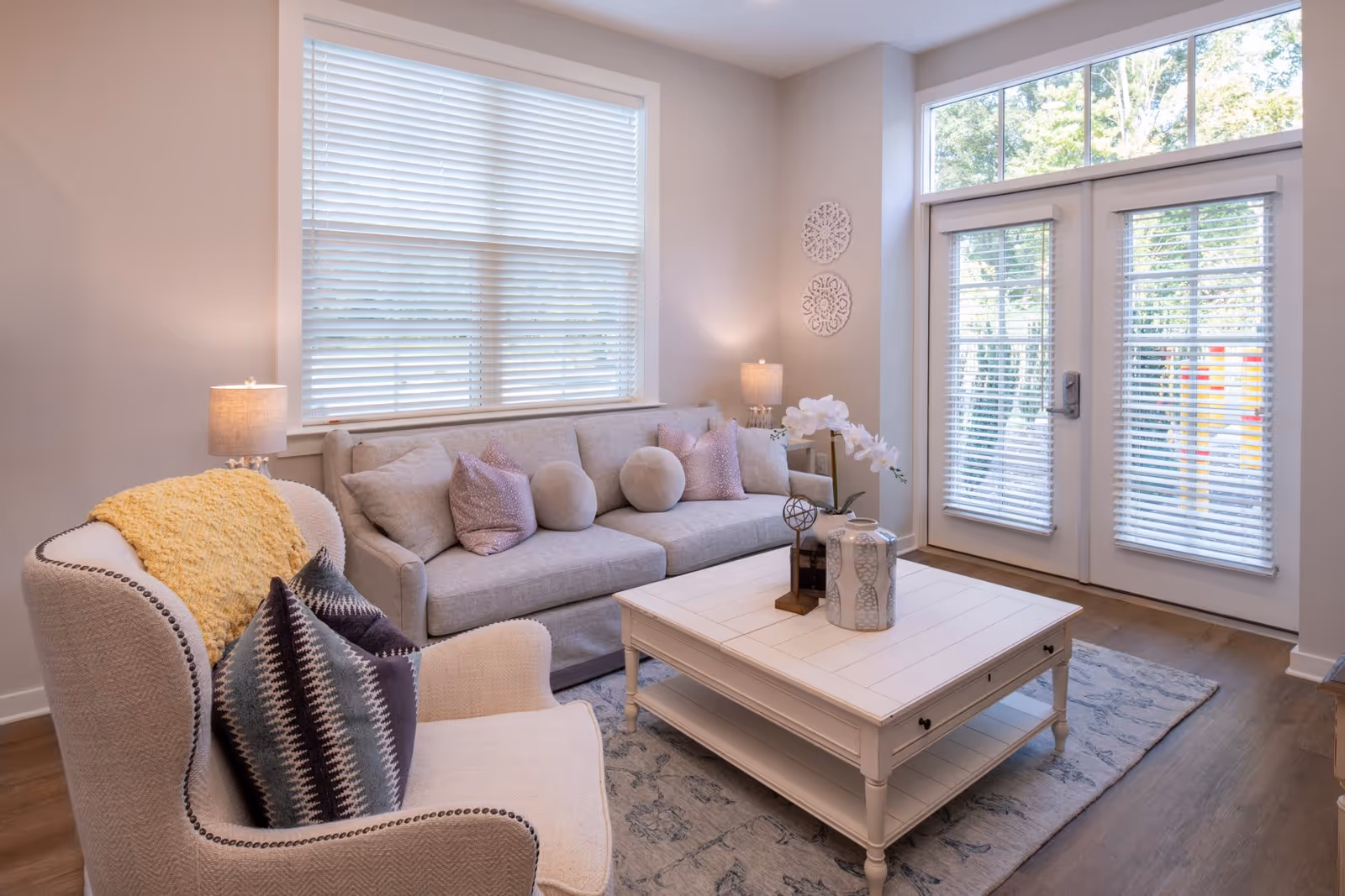 A cozy living room with a beige sofa adorned with round and square pillows, a cushioned armchair with patterned pillows and a yellow throw blanket, a white wooden coffee table with decorative items, two table lamps, large windows with blinds, and glass doors leading outside.