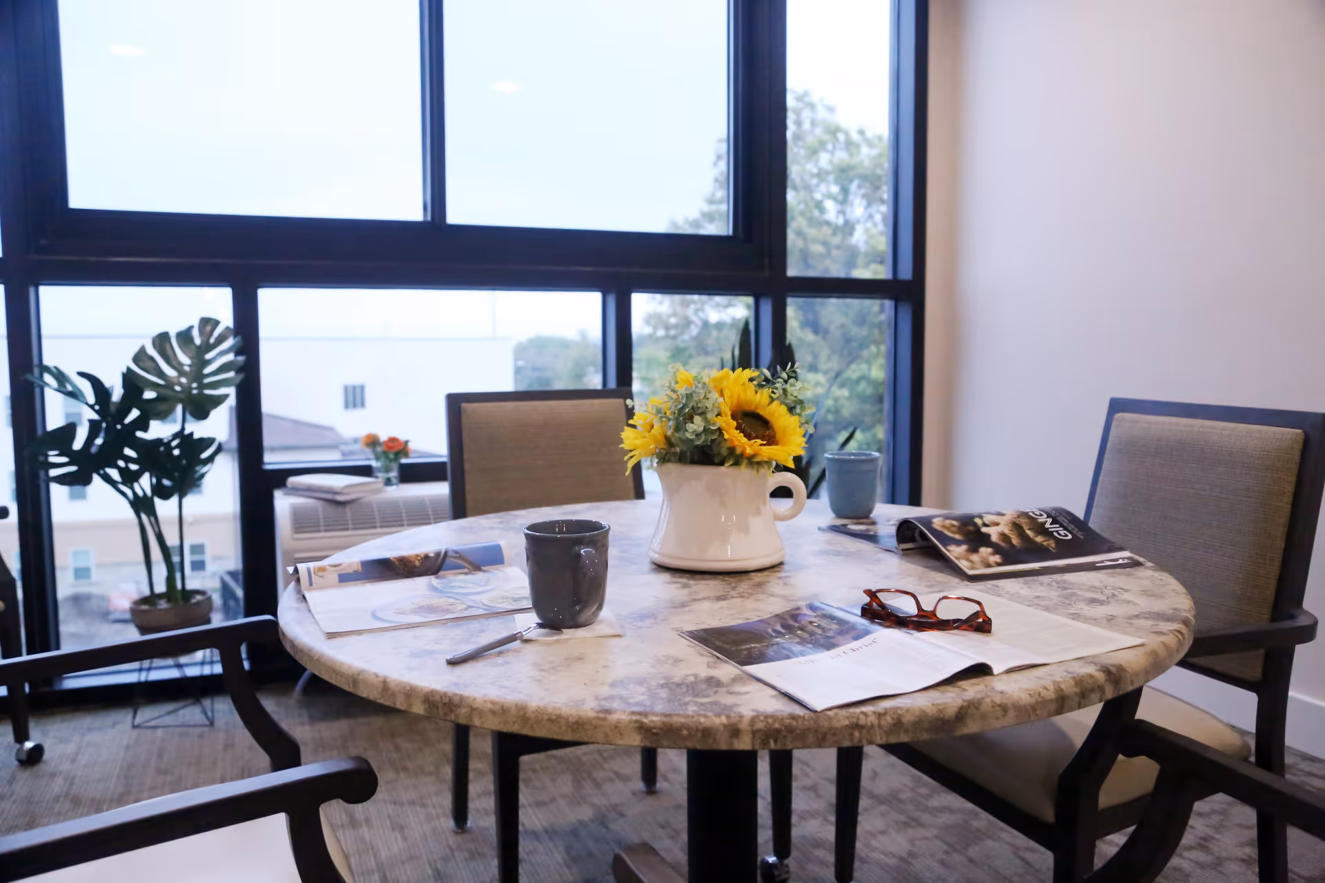 Round table with a vase of sunflowers, mugs, magazines and reading glasses in a bright room with large windows and chairs.