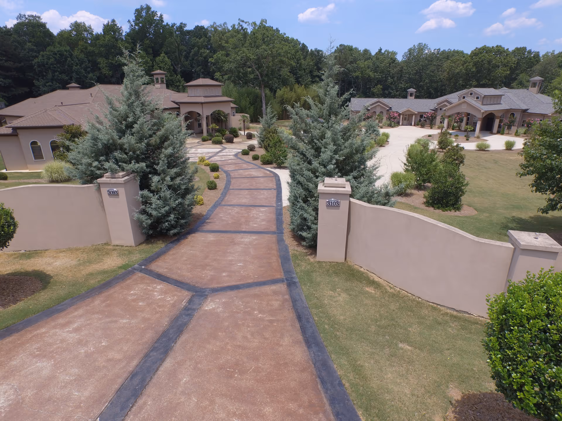 A wide paved driveway leading to a large, single-story senior living facility building surrounded by manicured lawns, shrubs, and trees under a blue sky with some clouds.