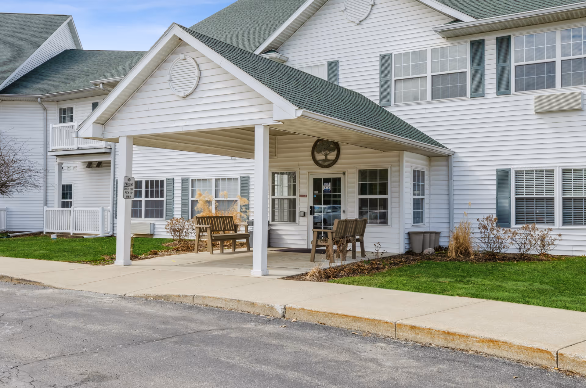 Covered driveway entrance of a white multi-story senior living building with benches and landscaping.