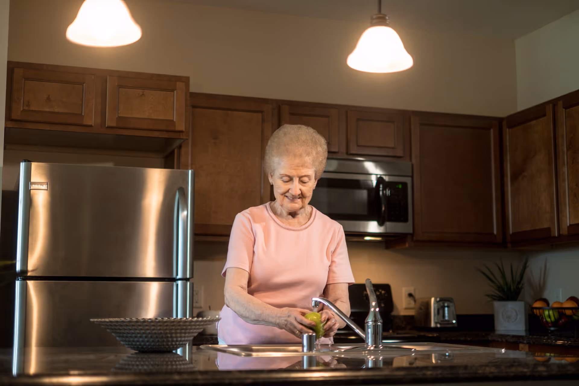 An elderly woman wearing a pink shirt is standing in a kitchen washing a green apple at the sink. The kitchen has wooden cabinets, a stainless steel refrigerator, a microwave, and a toaster. There are two pendant lights hanging from the ceiling and a decorative bowl on the counter.