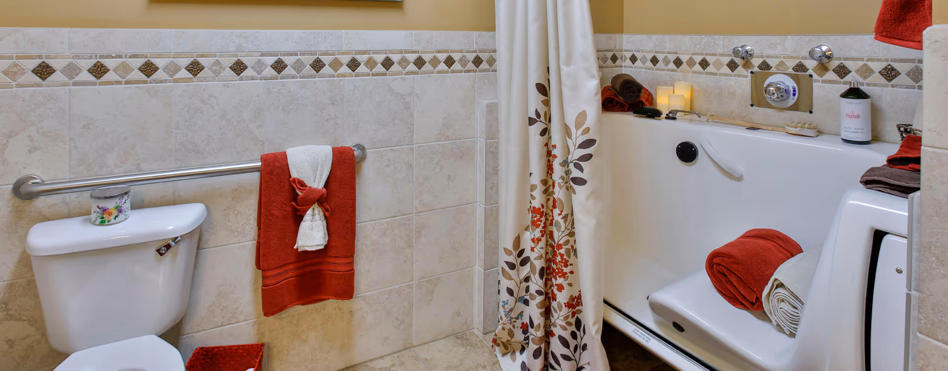 Accessible bathroom featuring a toilet with a grab bar, a walk-in bathtub with a floral shower curtain and folded towels, and tiled walls.