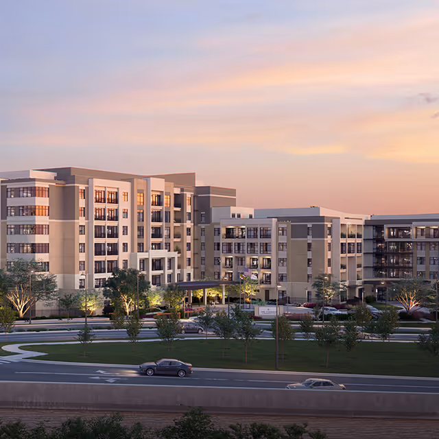 Exterior view of a large multi-story senior living facility building at dusk with cars on the road in front and landscaped greenery around the entrance.