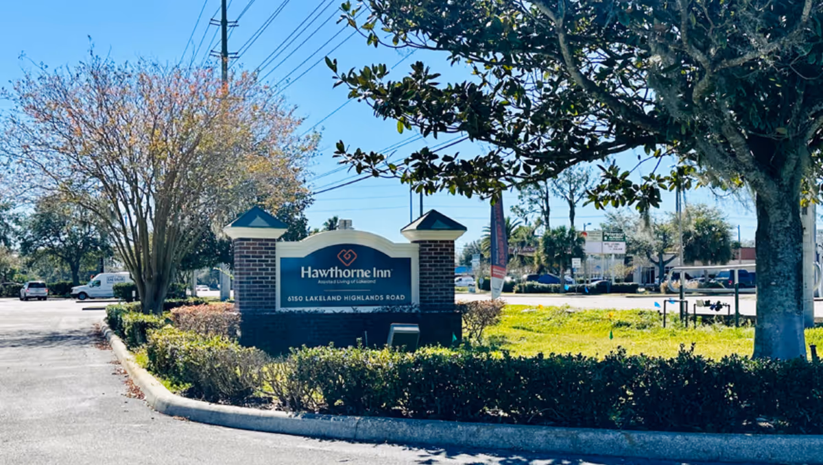 Outdoor view of the entrance sign for Hawthorne Inn Assisted Living of Lakeland, located at 6150 Lakeland Highlands Road, surrounded by trees, bushes, and a parking lot under a clear blue sky.