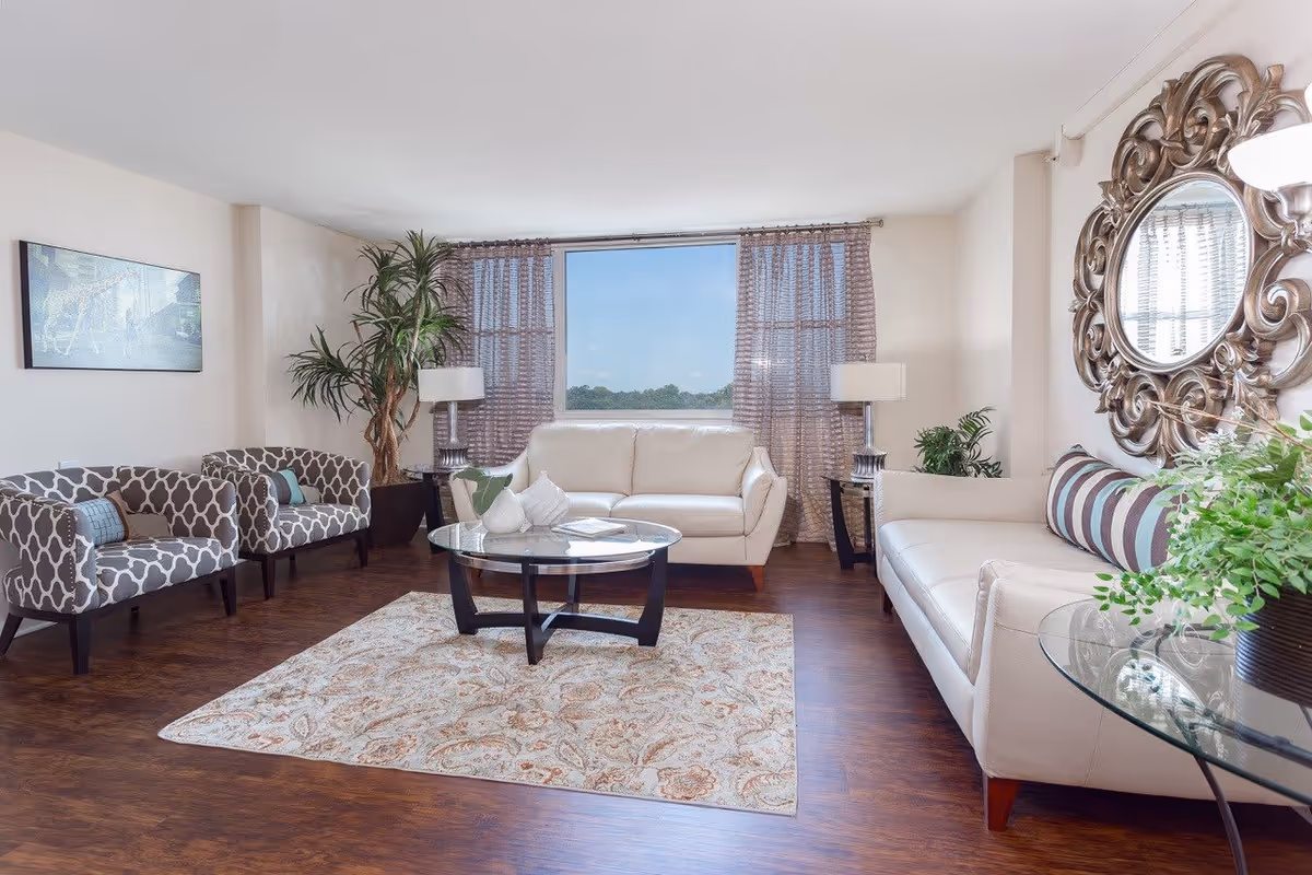 A bright and spacious living room with two white leather sofas, two patterned armchairs, a round glass coffee table on a decorative rug, a large ornate circular mirror on the wall, two table lamps on side tables, and a large window with sheer curtains showing a clear sky outside.