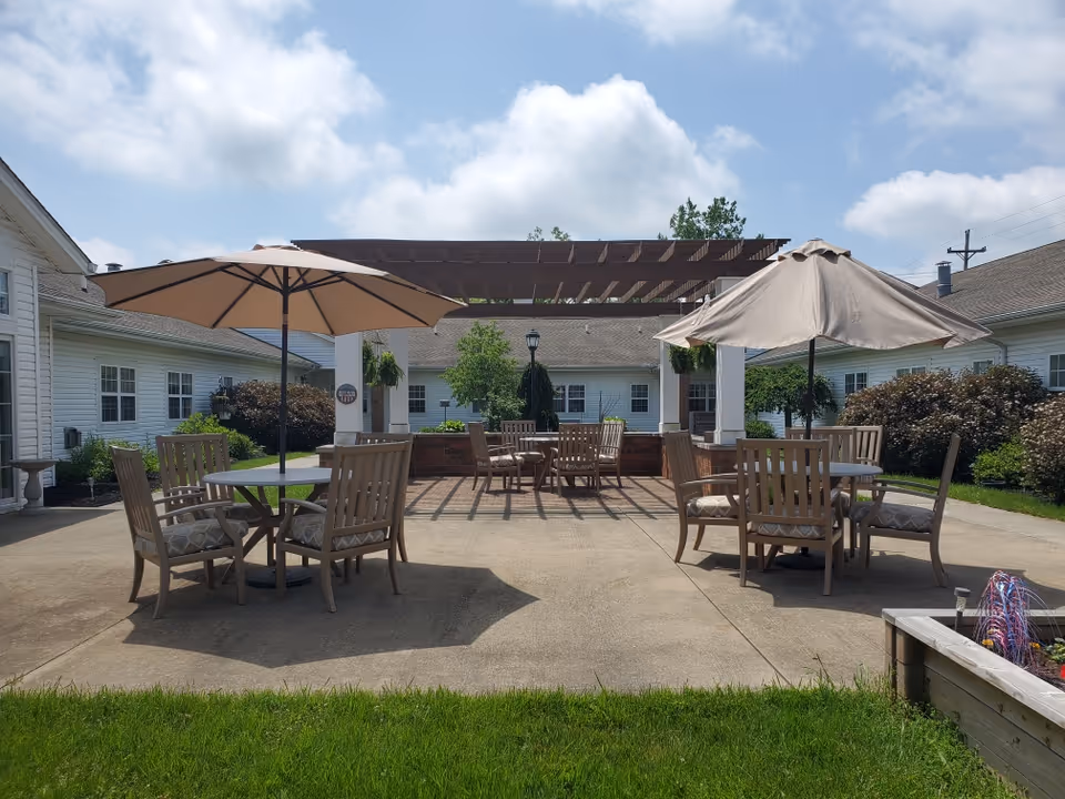 Outdoor patio area at a senior living facility with multiple round tables and wooden chairs, each table shaded by large beige umbrellas. The patio is surrounded by white buildings with windows, greenery, and a pergola structure in the background under a partly cloudy sky.