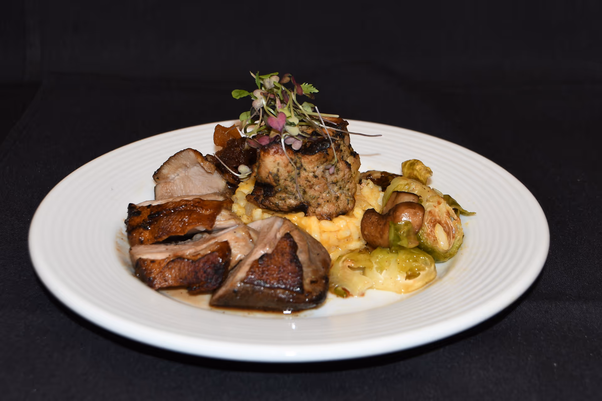 A white plate with sliced roasted meat, a herb-crusted meat roulade on mashed potatoes, and Brussels sprouts garnished with microgreens.
