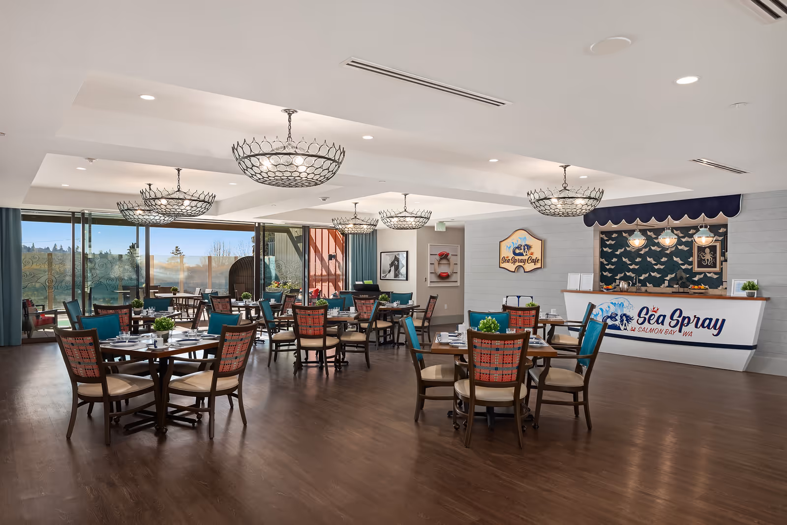 A spacious dining room with multiple wooden tables and chairs arranged neatly. Each table is set with plates, cups, and small green plants as centerpieces. The room features large windows allowing natural light to enter, and a counter area labeled 'Sea Spray Cafe' with decorative lighting and a marine-themed backdrop. The floor is wooden, and the ceiling has multiple modern chandeliers.