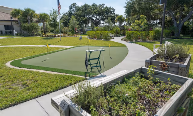 Outdoor garden area at Inspired Living Ocoee featuring a small putting green with three yellow flags, a chair, raised garden beds with plants, a paved walkway, and trees and shrubs in the background under a clear sky.