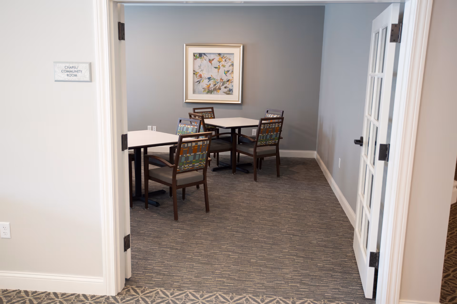 View through an open doorway into a small room with two square tables and four chairs with patterned backs. The room has gray walls, carpeted floor, and a framed artwork featuring birds on the far wall. A sign on the left side of the doorway reads 'CHAPEL/COMMUNITY ROOM'.