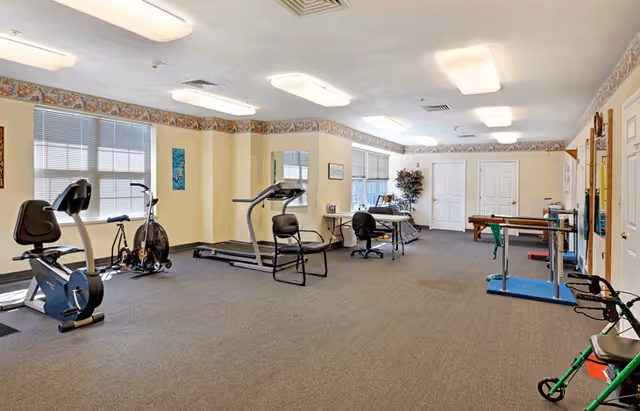 A spacious senior living exercise room with various fitness equipment including a stationary bike, treadmill, and exercise bike. The room has beige walls with a floral border near the ceiling, large windows letting in natural light, fluorescent ceiling lights, and a carpeted floor. There are chairs and tables along the walls, and a walker is visible in the foreground.
