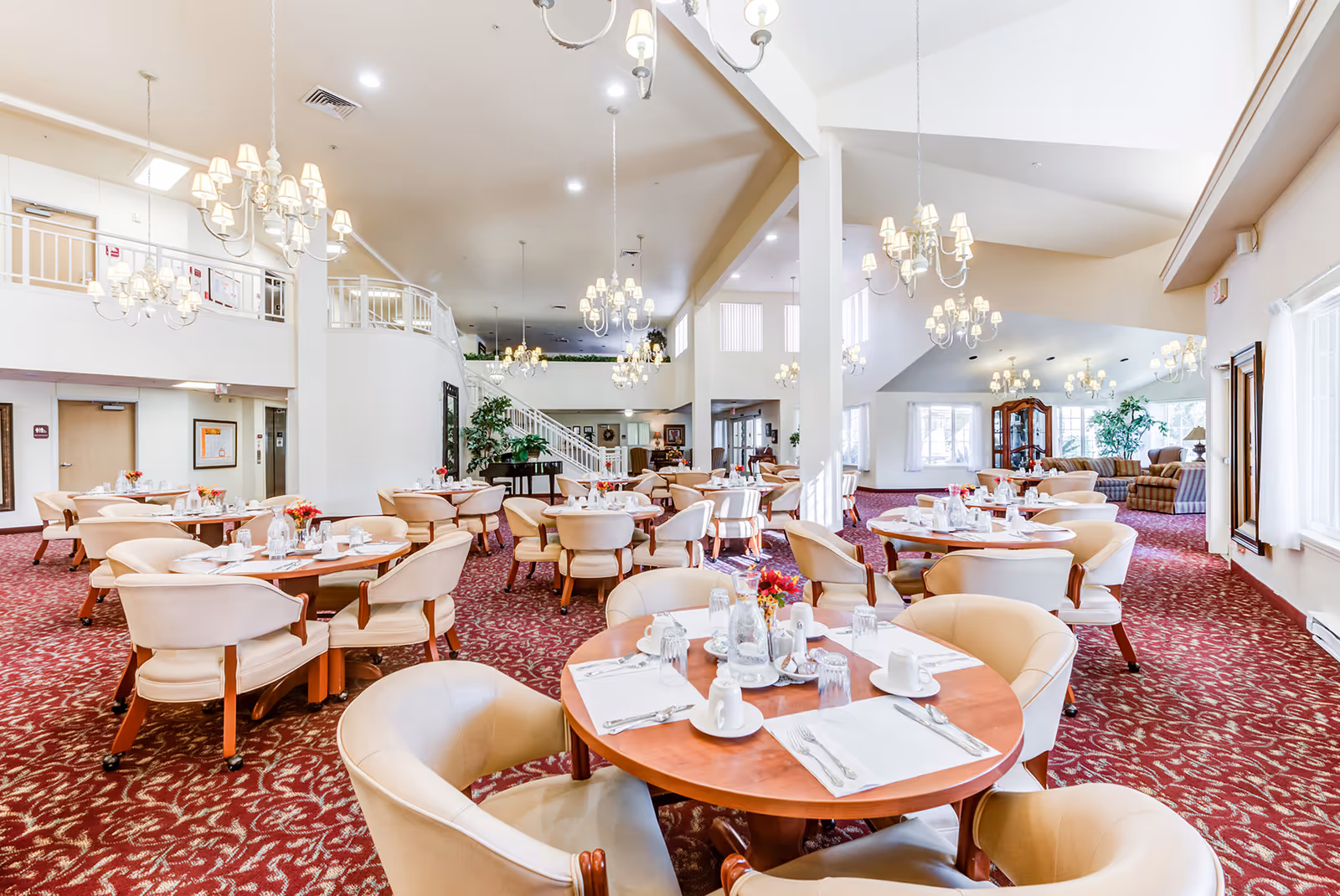 Spacious assisted-living dining room with round tables set for meals, beige chairs, chandeliers, and a red patterned carpet.