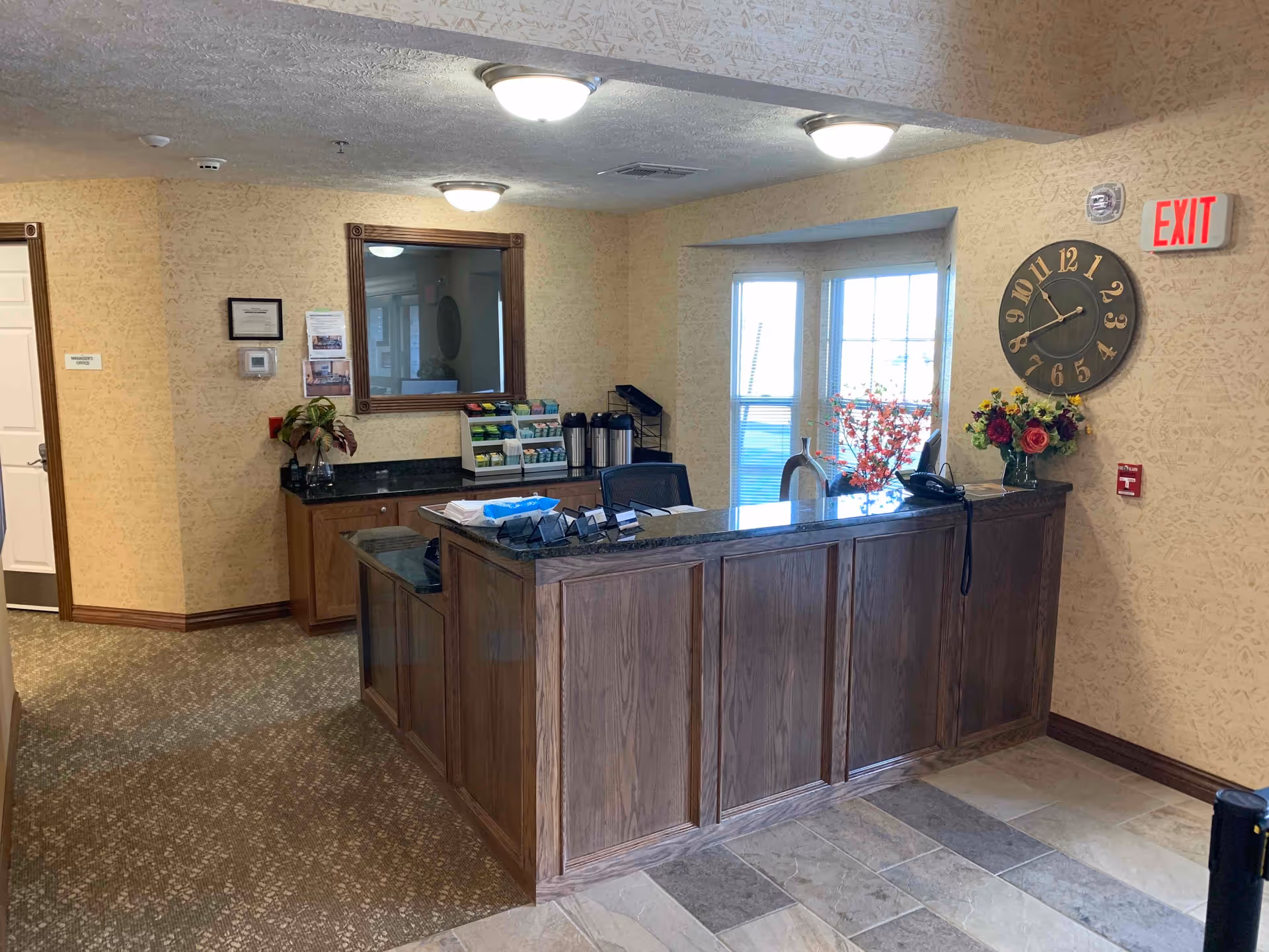 Reception area with a wooden front desk, a large wall clock, an exit sign, a telephone, and a vase with flowers. Behind the desk is a counter with coffee dispensers, a mirror, and informational pamphlets. The walls are light-colored with a subtle pattern, and the floor is a mix of carpet and tile.