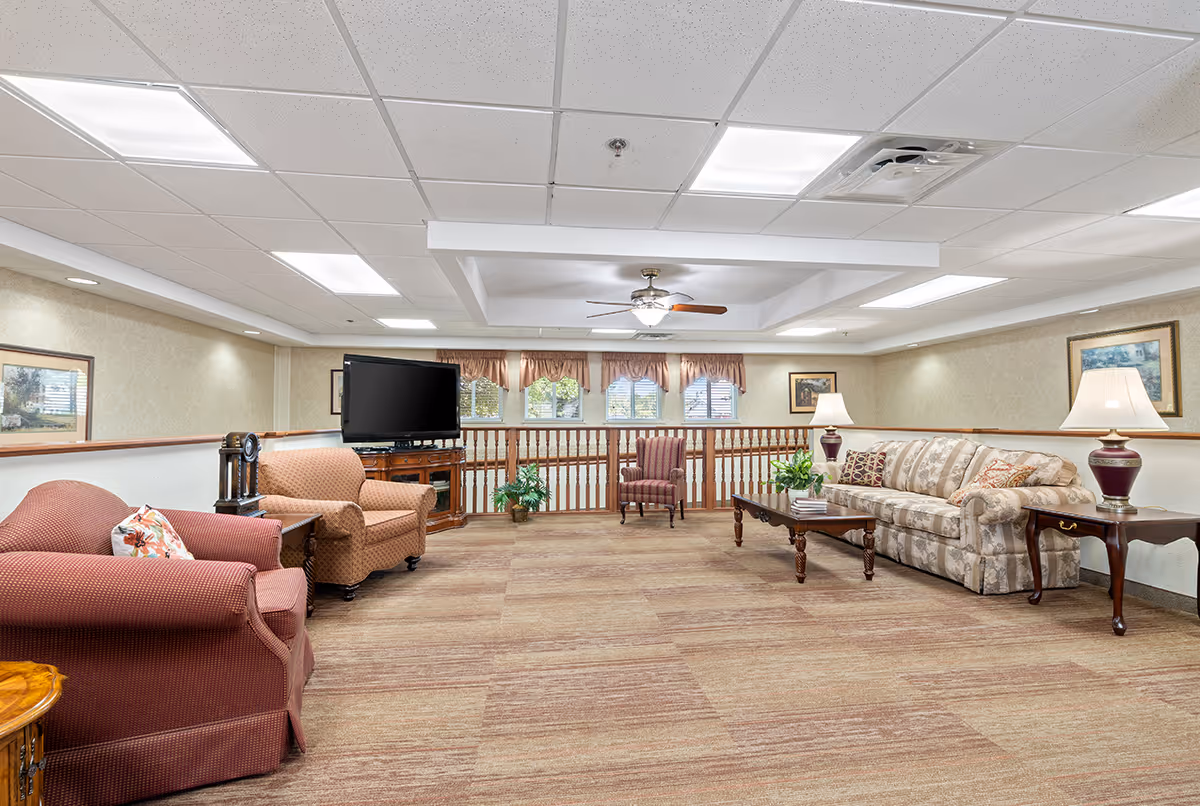 A spacious living room area with beige walls and carpeted floor. The room features a patterned sofa with decorative pillows, two armchairs, a wooden coffee table with plants and books, side tables with lamps, a flat-screen TV on a wooden stand, and framed artwork on the walls. There are windows with valances and a ceiling fan with lights in the center of the ceiling.