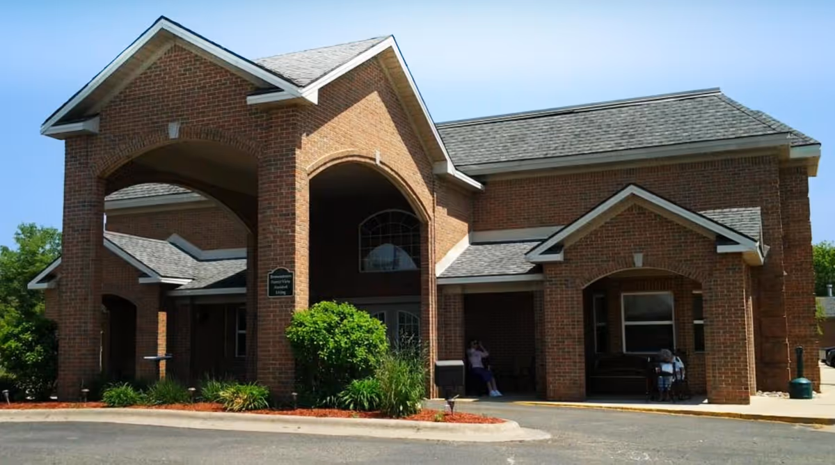 Brick exterior and covered entrance/porte-cochère of an assisted living building with landscaping and two people seated near the entry.