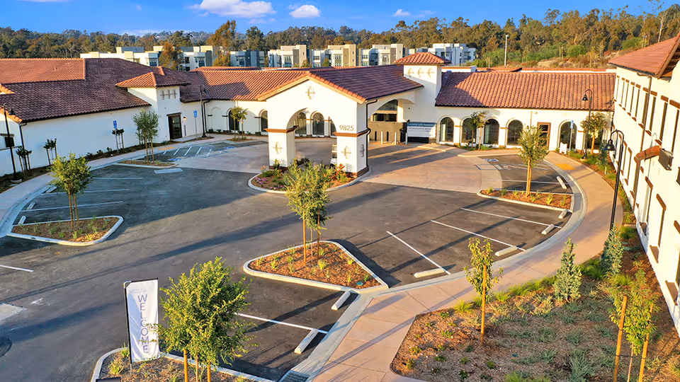 Exterior view of Ridgeview Health Center showing a large parking lot with marked parking spaces and landscaped islands with small trees. The building has white walls and red-tiled roofs with arched windows and a covered entrance area. The surrounding area includes trees and a clear blue sky.