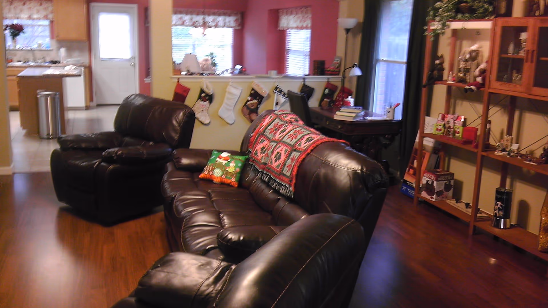 Cozy living room with dark brown leather sofas, a colorful quilt and a Christmas-themed pillow on the couch. The room has wooden flooring and a shelf with various decorative items. In the background, there is a kitchen area with wooden cabinets and a white door. Christmas stockings are hung on a half wall separating the living room and kitchen.