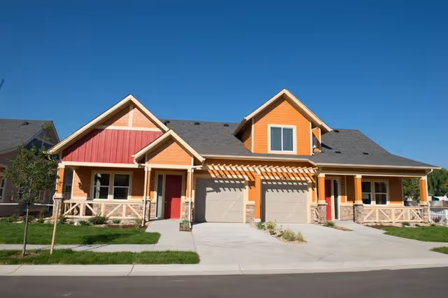 Exterior view of a modern duplex house with orange and red siding, two garages, and a well-maintained lawn under a clear blue sky.