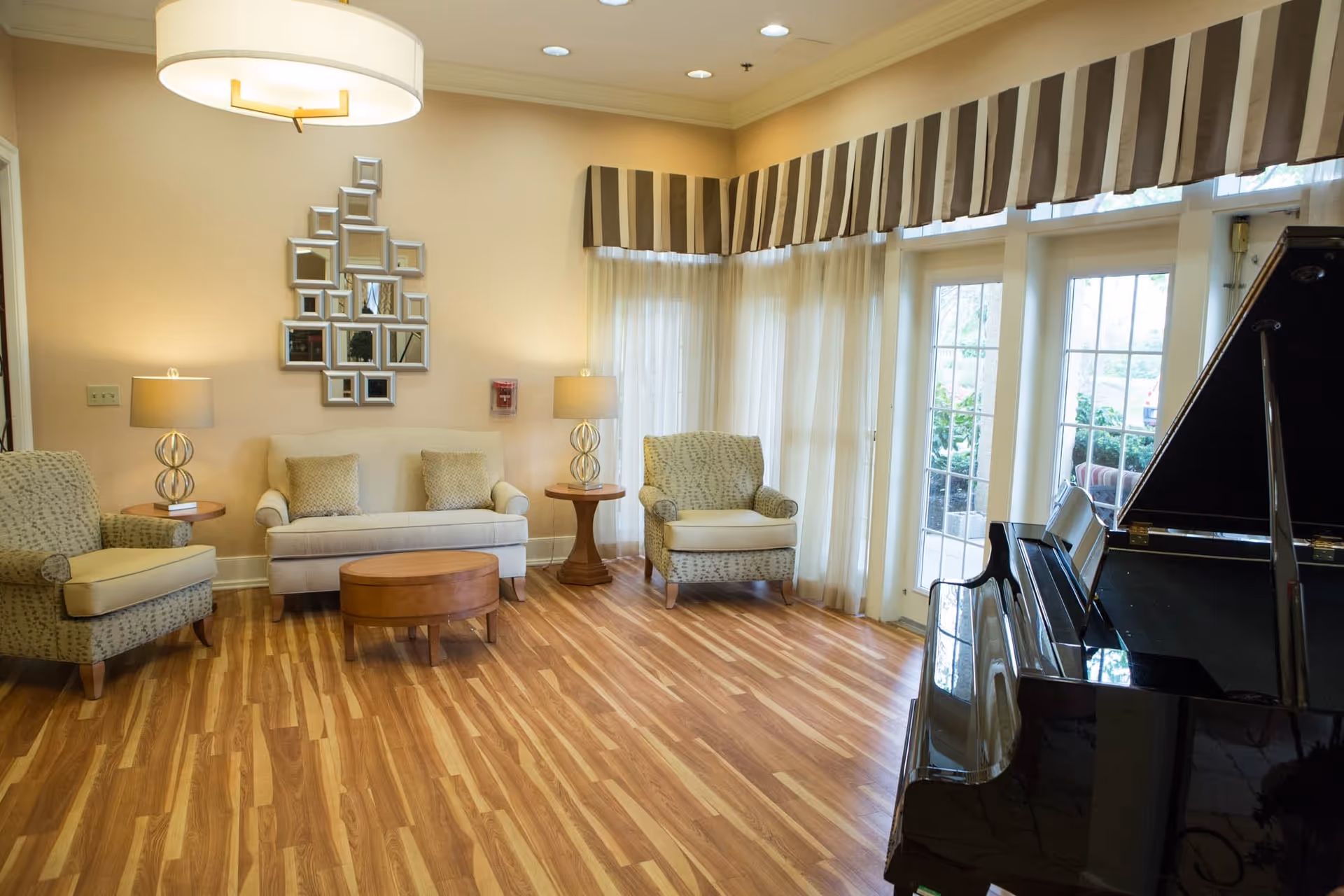 A cozy living room area with wooden flooring, featuring a beige sofa with two cushions, two patterned armchairs, two side tables with lamps, a round wooden coffee table, a decorative mirror on the wall, large windows with sheer curtains and striped valances, and a black grand piano on the right side.
