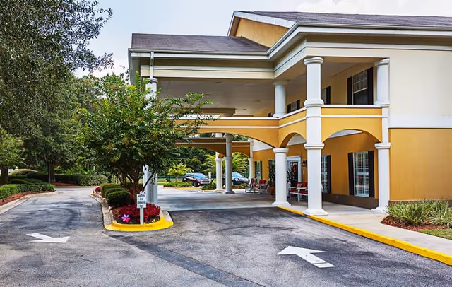 Covered porte-cochère and driveway at the front of a two-story yellow building with columns and landscaped surroundings.