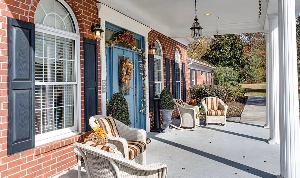 Front porch of a brick building with a blue door decorated with a fall wreath and garland. The porch has white wicker chairs with striped cushions, potted plants, and black shutters on the windows. There are white columns supporting the porch roof and outdoor lantern-style lights.
