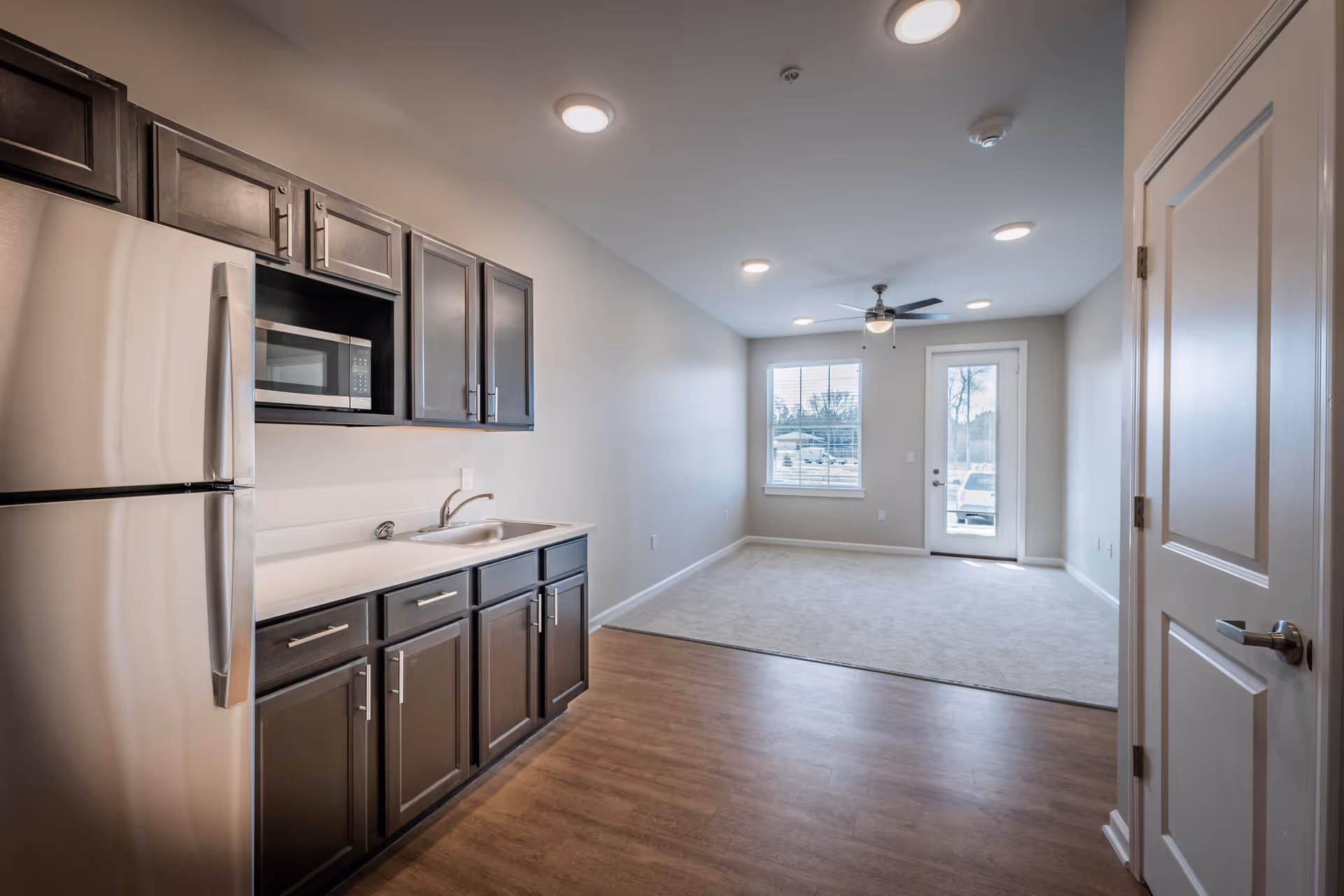 Interior view of a senior living facility unit showing a kitchen area with dark wood cabinets, a stainless steel refrigerator, microwave, and sink on the left side. The kitchen opens into a carpeted living area with a ceiling fan, a window, and a glass door leading outside.