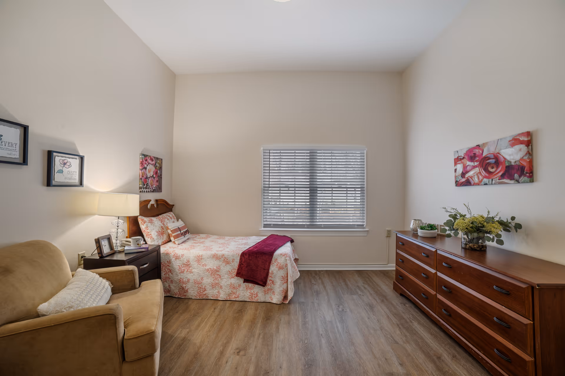 A tidy bedroom with a twin bed dressed in floral bedding, an armchair, nightstand with lamp, and a wooden dresser beneath a window with blinds.