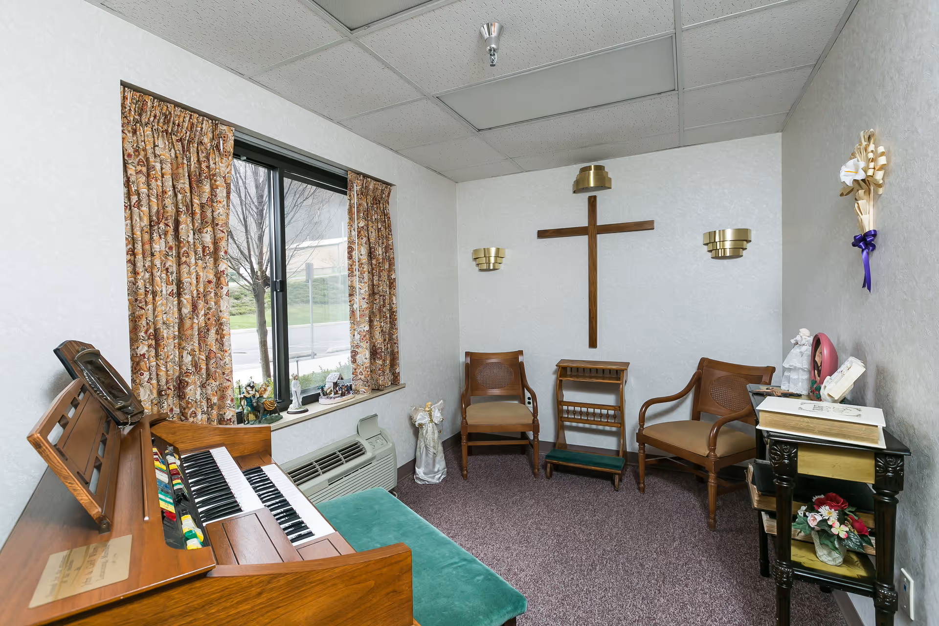 A small room with a wooden organ and green cushioned bench on the left, two wooden chairs with armrests, a wooden cross mounted on the wall, and a small table with decorative items including flowers and a book. The room has floral curtains on the window and beige walls with carpeted floor.