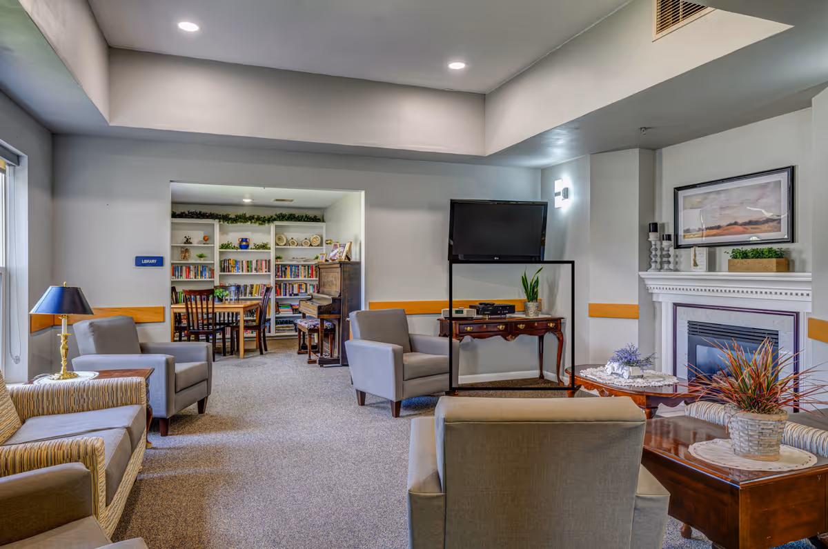 A cozy living room area with several armchairs and sofas arranged around a wooden coffee table with a plant on it. There is a fireplace with decorative items and a framed picture above it. A flat-screen TV is mounted on the wall above a wooden console table with a plant and electronic devices. In the background, there is a small library area with bookshelves filled with books, a piano, and a table with chairs. The room has soft lighting and a carpeted floor.